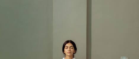 A person meditating in a minimalist room with a plain wall, a cushioned bench, and two candle stands.