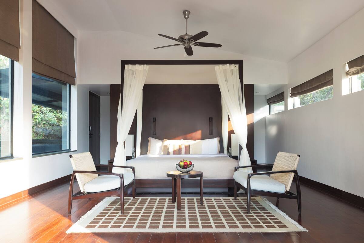 A spacious bedroom with a large bed, two chairs, a small table with a fruit bowl, and a ceiling fan.