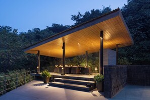 A modern outdoor dining area with a wooden roof and steps leading up to it, surrounded by greenery. - Dharana at Shillim - Luxury Clinical Wellbeing Retreat (Mawal)