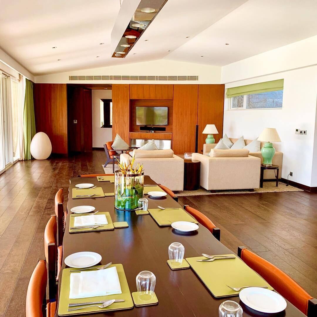 A modern dining area with a long table set for a meal, a television in the background, and a cozy seating area with sofas and lamps.
