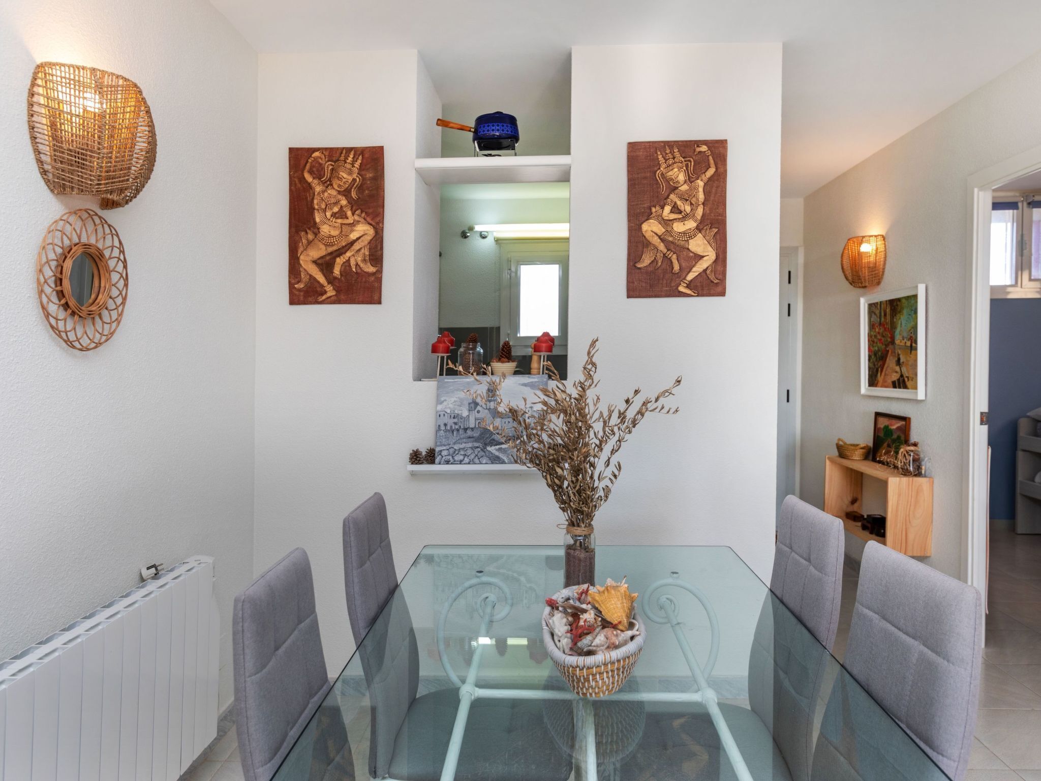 A dining area with a glass table, grey chairs, and a vase with dried plants. There are two wall-mounted artworks and a wicker lamp.