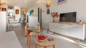 A modern living room with a sofa, two small round tables with fruit bowls and wine glasses, a TV on a wooden stand, and artwork on the walls. - Edificio Luxor II by Interhome (Miami Platja)