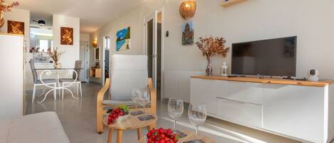A modern living room with a sofa, two small round tables with fruit bowls and wine glasses, a TV on a wooden stand, and artwork on the walls.