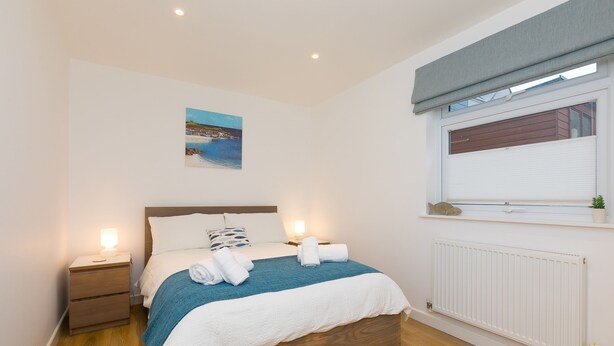 Ein ordentlich gemachtes Bett mit blauer Decke, ein Nachttisch mit Lampe, ein Fenster mit Blick auf ein Gebäude und ein an der Wand hängendes Bild.