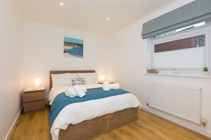 Uma cama bem arrumada com um cobertor azul, uma mesa de cabeceira com abajur, uma janela com vista para um edifício e um quadro pendurado na parede.