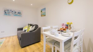 Ein modernes Wohnzimmer mit grauem Sofa, weißem Esstisch und Stühlen. An den Wänden sind Kunstwerke, eine Uhr und ein kleiner Tisch mit Speisen und Getränken zu sehen.