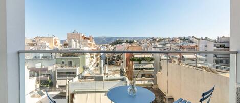 A balcony with a table and chairs, overlooking a cityscape with buildings and a distant mountain range.