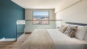 A bedroom with a bed, a window with a city view, and a glass railing.