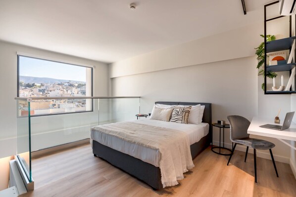 A modern bedroom with a large bed, a desk, and a laptop. There is a view of a cityscape through a large window.