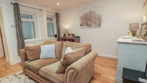 A living room with a sofa, a small table, a painting, and a window with curtains. - Rosalin Cottage (Irvine)