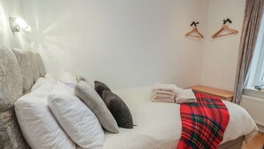 A bedroom with a bed, pillows, a blanket, a wall-mounted light, and a window with curtains. - Rosalin Cottage (Irvine)