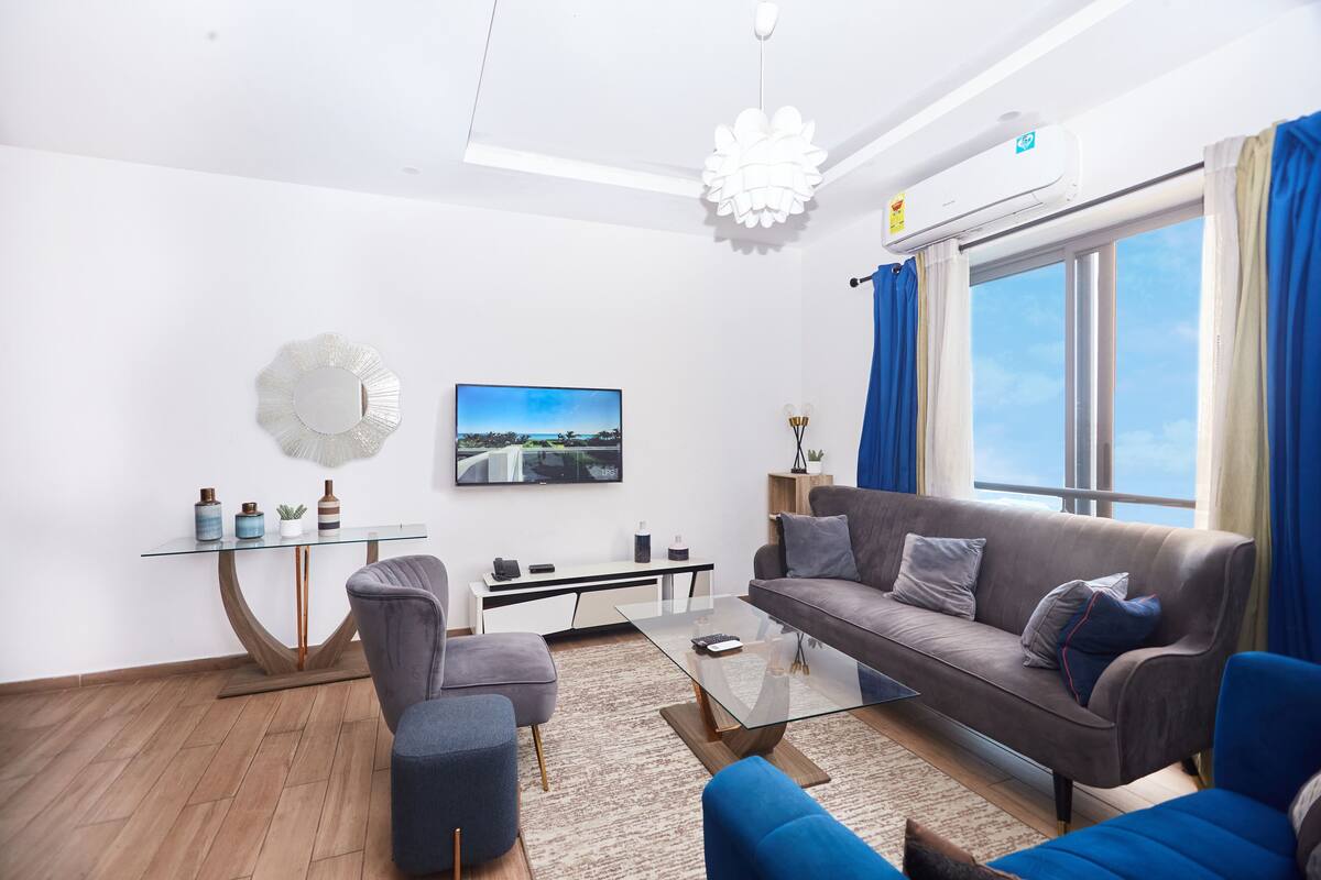 A modern living room with a grey sofa, blue armchairs, a glass coffee table, a small side table, a mounted TV, and a large window with curtains.