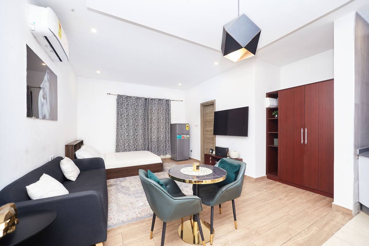 A modern hotel room with a sofa, two chairs, a small round table, a TV, a wardrobe, and a bed.