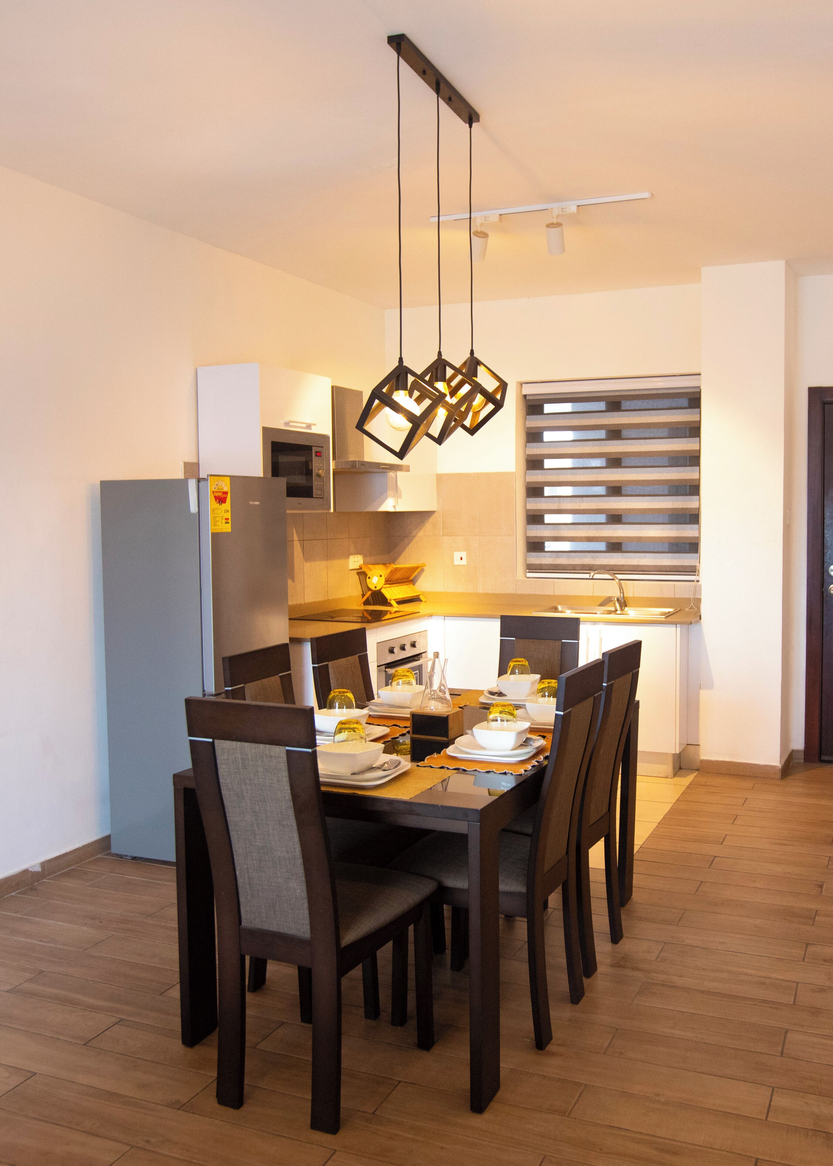 Een moderne eethoek met een houten tafel en stoelen, een keuken op de achtergrond en hanglampen.