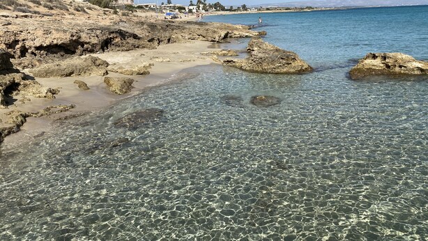 Una playa rocosa con aguas cristalinas y edificios al fondo.