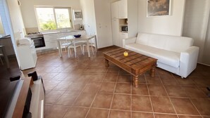Un amplio salón con un sofá blanco, una mesa de centro de madera y una mesa de comedor con sillas. Se ve una cocina con armarios blancos y una ventana al fondo.