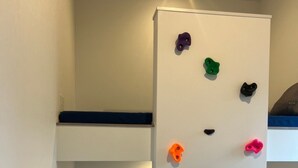 A room with a bunk bed, a climbing wall with colorful holds, and a blue mattress on the lower bunk. - BBQ (Mihama)