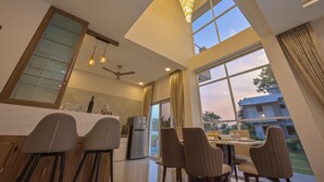 A modern dining area with a high ceiling, large windows, and a chandelier. - Vivanta Villa (Mawal)