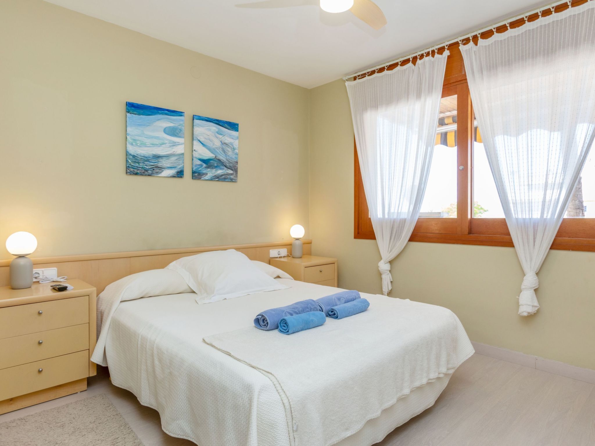 A bedroom with a bed, two bedside tables, a nightstand, a ceiling fan, a window with curtains, and two wall-mounted paintings.
