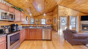 A spacious kitchen with wooden cabinets, stainless steel appliances, and a central island. There is a dining area with a sofa and a fireplace. - Bear Cove by AvantStay | Hot Tub + Pool Table | In the Heart of Pigeon Forge (Pigeon Forge)