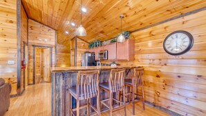 A wooden cabin interior with a kitchen area, wooden bar stools, and a clock on the wall. - Bear Cove by AvantStay | Hot Tub + Pool Table | In the Heart of Pigeon Forge (Pigeon Forge)