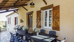 Une terrasse extérieure avec un sol en pierre, une table noire entourée de chaises et des volets en bois aux fenêtres.