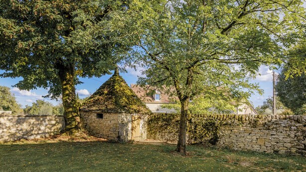 Une construction en pierre surmontée d’un toit conique, entourée d’un mur de pierre et d’arbres.