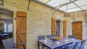 Un bâtiment rustique en pierre, doté d’une grande table en bois dressée pour le repas, entourée de chaises en osier.