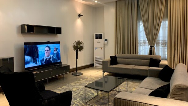 A modern living room with a flat-screen TV, a ceiling fan, a sofa, and a coffee table. - De James Hotels and Suites (Abuja)
