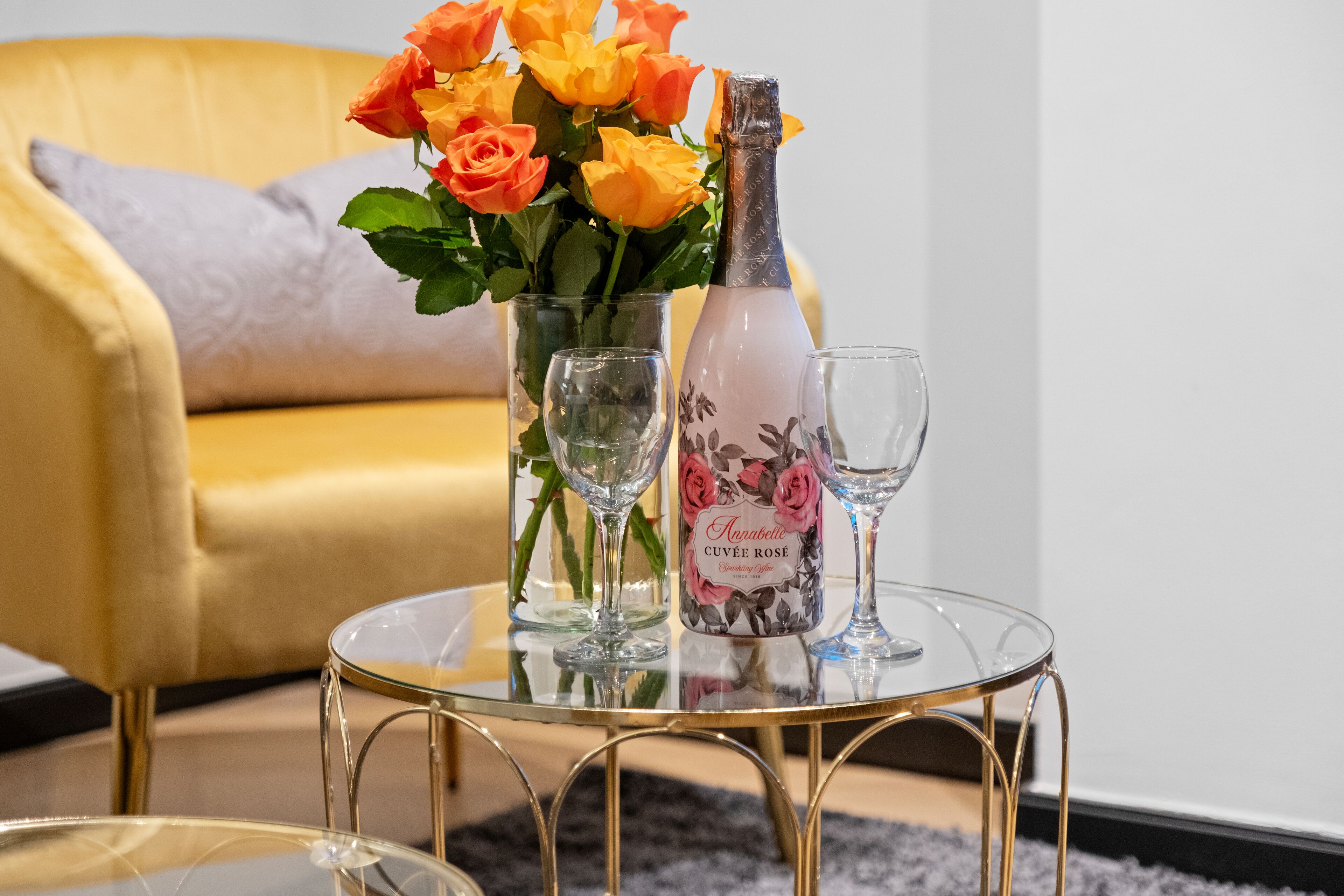 A glass table with a bottle of Anabellé Cuvée Rose Champagne, a vase with orange roses, and two wine glasses.