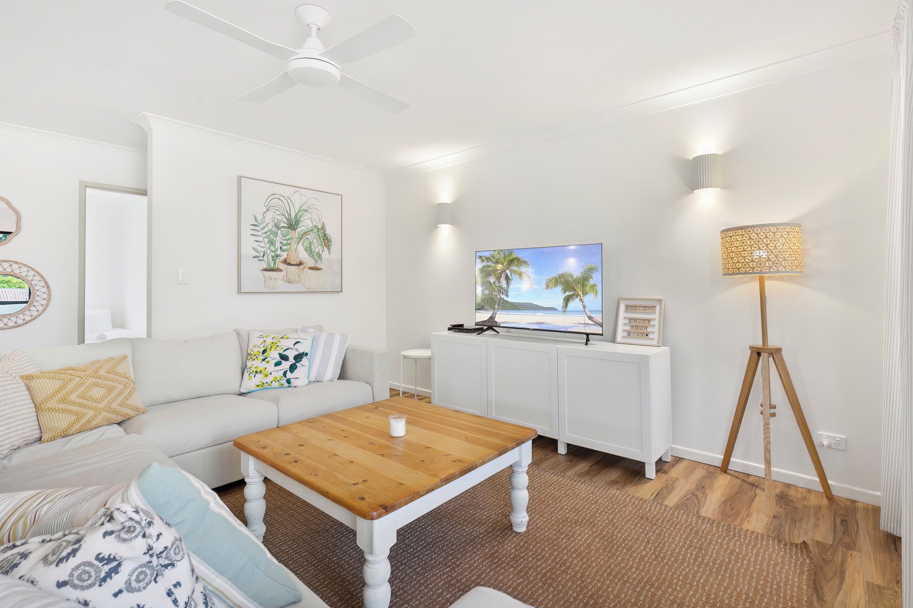 Uma sala de estar moderna com um sofá branco, uma mesa de centro de madeira, uma televisão em um móvel branco, um abajur e um ventilador de teto.