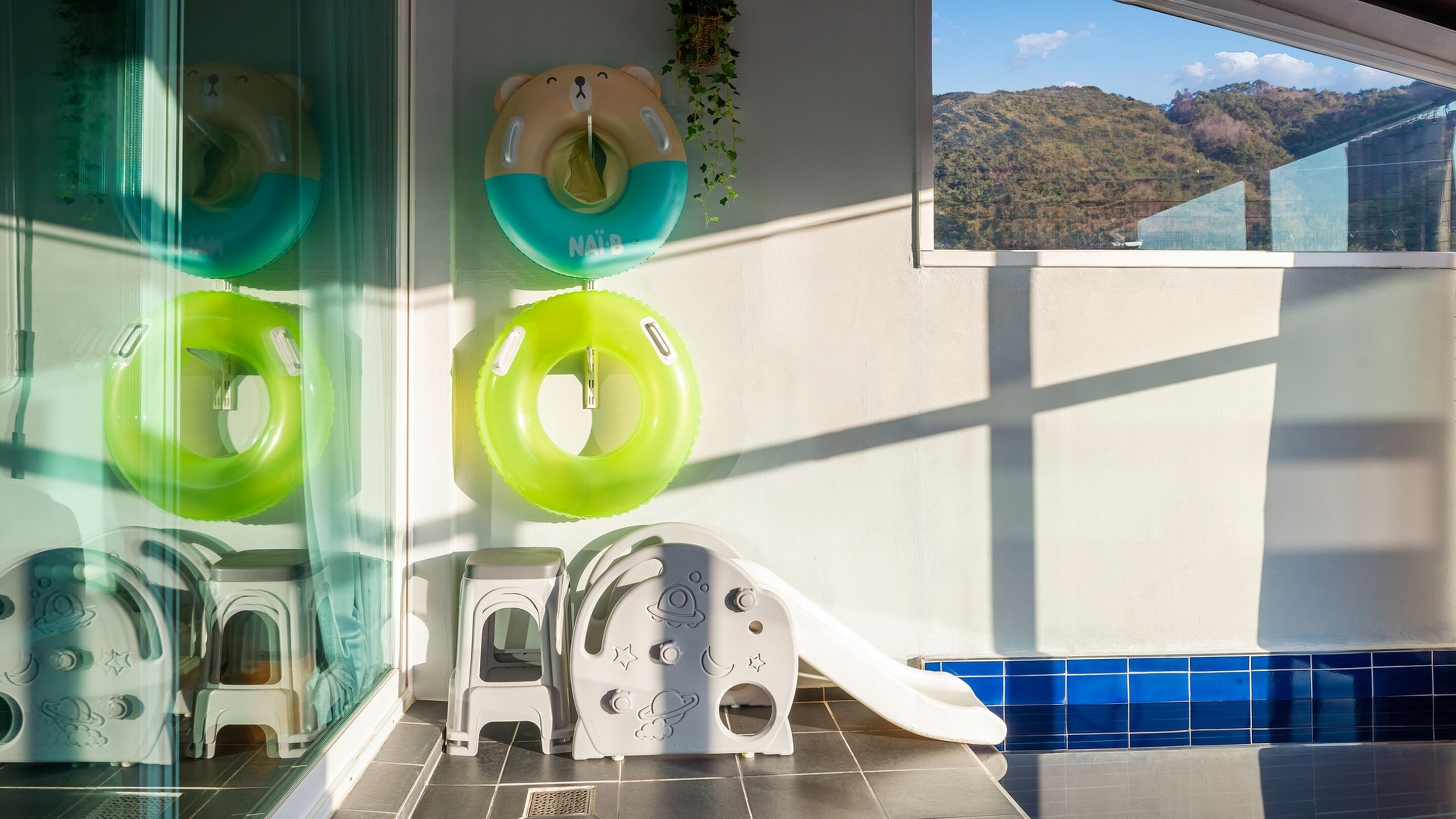 A children's bathroom with a slide, two green and one beige inflatable pool float, and a view of a mountain through a window.