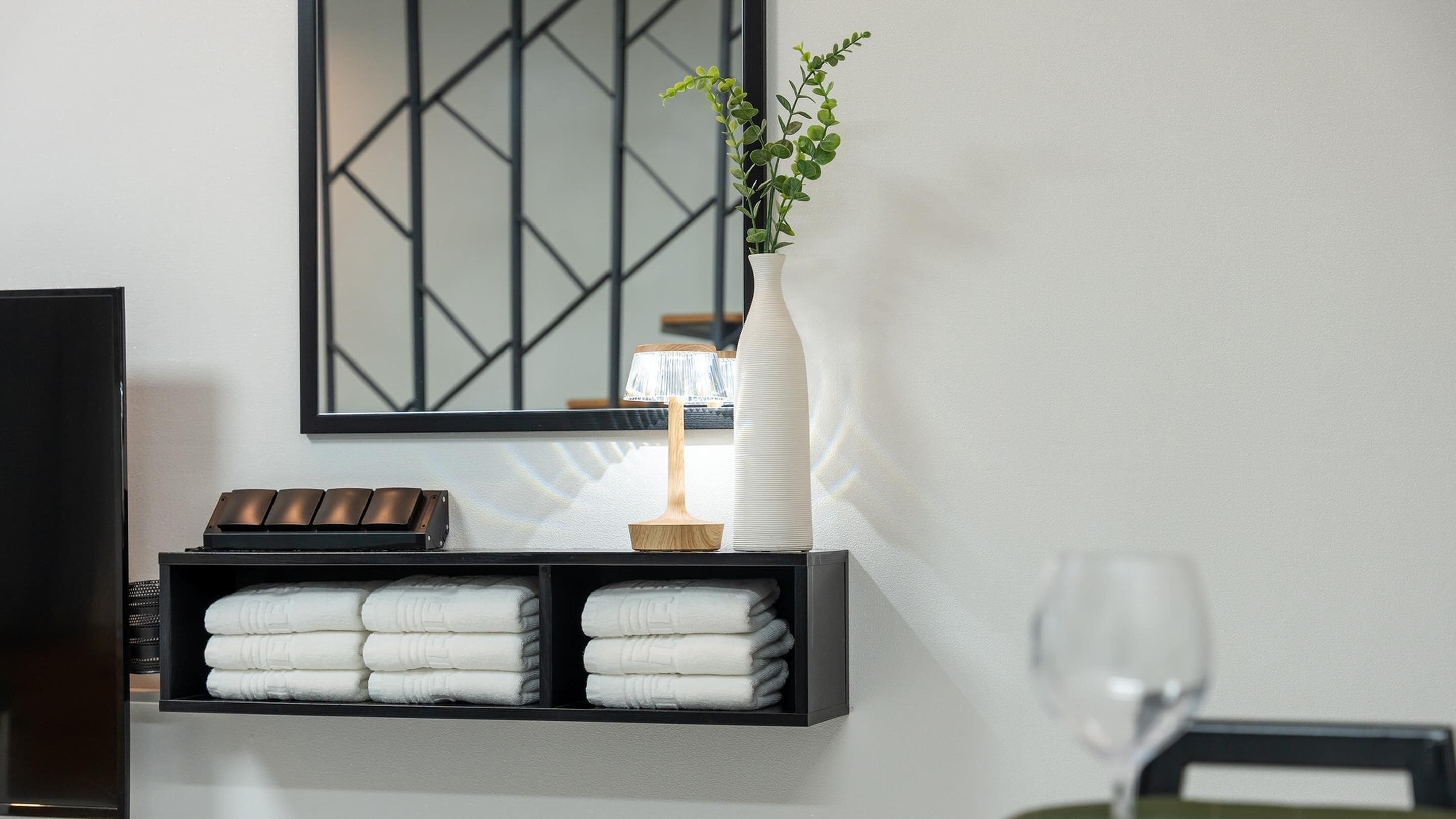 A bathroom with a shelf holding towels, a vase with a plant, and a mirror with a geometric pattern.