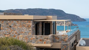 Un bâtiment moderne avec une façade en pierre, un balcon doté d’une rambarde en verre, et une vue sur la mer et une montagne lointaine.