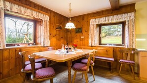 Interior de una cabaña de madera con una mesa de comedor, cuatro sillas y dos ventanas con cortinas.
