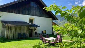 Un chalé rústico con techo inclinado, fachada de madera y frontis blanco. Cuenta con un patio con mobiliario exterior y vistas a un paisaje montañoso.