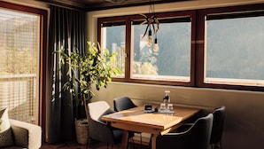 Comedor con mesa de madera, sillas, planta en maceta y ventanas grandes con cortinas.