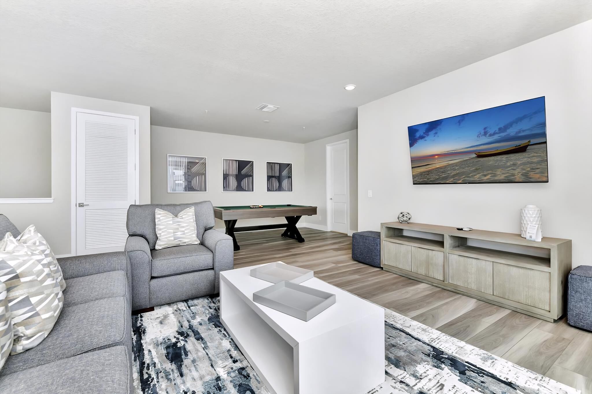A modern living room with a grey sofa, a white coffee table, a pool table, and a painting of a beach scene.