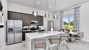 A modern kitchen with stainless steel appliances, a central island with bar stools, pendant lighting, and a view of a garden through large windows. - Inviting Condo Nook near Disney with Resort & Waterpark Access - B44-239 (Kissimmee)