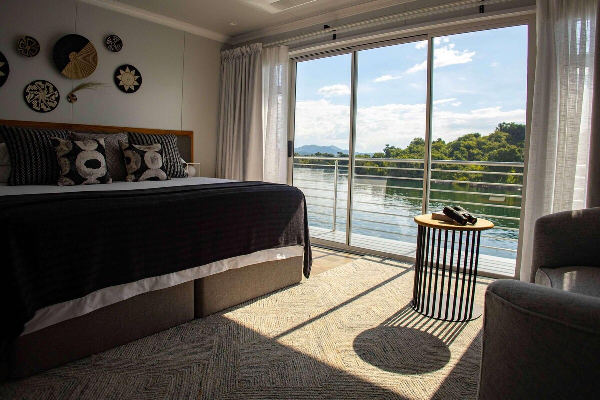 A bedroom with a large bed, a view of a lake, and a balcony with a table and chair.
