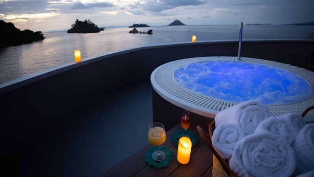 Un bain à remous avec vue sur la mer et les îles, une bougie et des serviettes sur une table en bois.