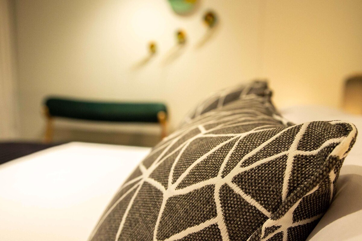 Een detail van een gestreept kussen op een bed, met een wazige achtergrond van een groene bank en decoratieve wandelementen.