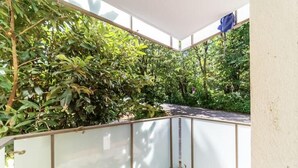 A covered balcony with a glass railing, surrounded by lush greenery.