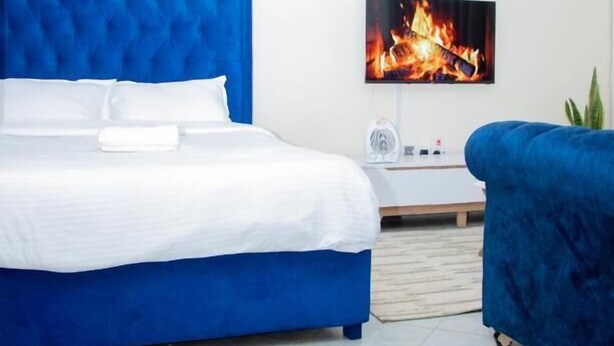 Une chambre moderne avec un lit recouvert d’un tissu bleu, une table de chevet blanche et un téléviseur fixé au mur qui affiche une image de feu.
