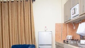 Une cuisine compacte équipée d’un réfrigérateur, d’un four à micro-ondes et d’un grille-pain. Un canapé bleu est placé contre le mur, et un rideau est partiellement tiré.