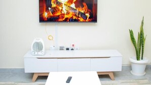 Un salon moderne avec un téléviseur à écran plat fixé au mur, un meuble de rangement blanc en dessous, une petite table avec une télécommande et une plante en pot placée sur le côté.