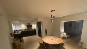 Eine moderne Küche mit dunklen Schränken und einer zentralen Kochinsel, ein Essbereich mit einem runden Holztisch und Stühlen, und eine gemütliche Sitzecke mit Lampe und Kissen.