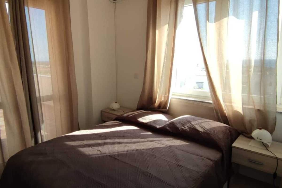 Quarto com cama, mesa de cabeceira, abajur e cortinas translúcidas.
