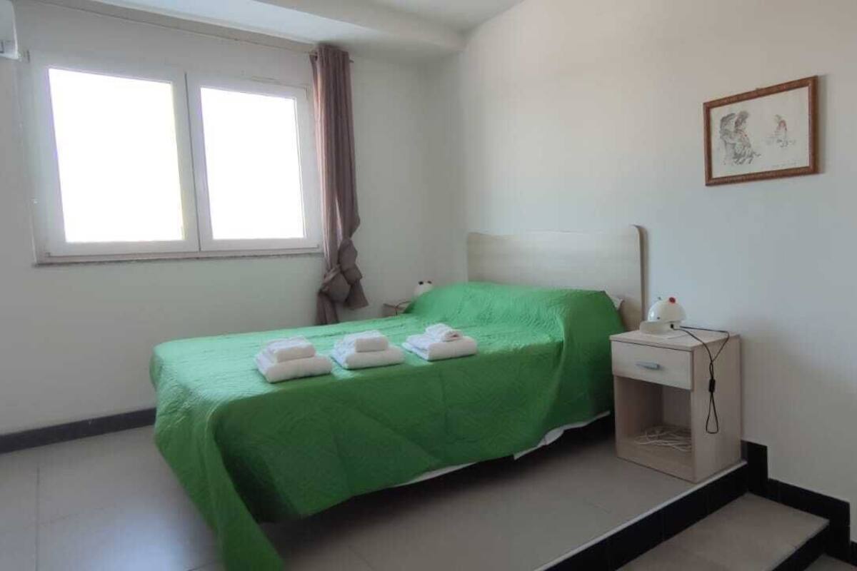 Quarto com cama, mesa de cabeceira, janela, porta e banheiro com pia.