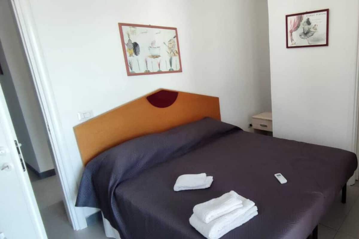 Quarto de hotel com cama, duas toalhas, mesa de cabeceira e uma pequena mesa. Há duas molduras com quadros na parede.